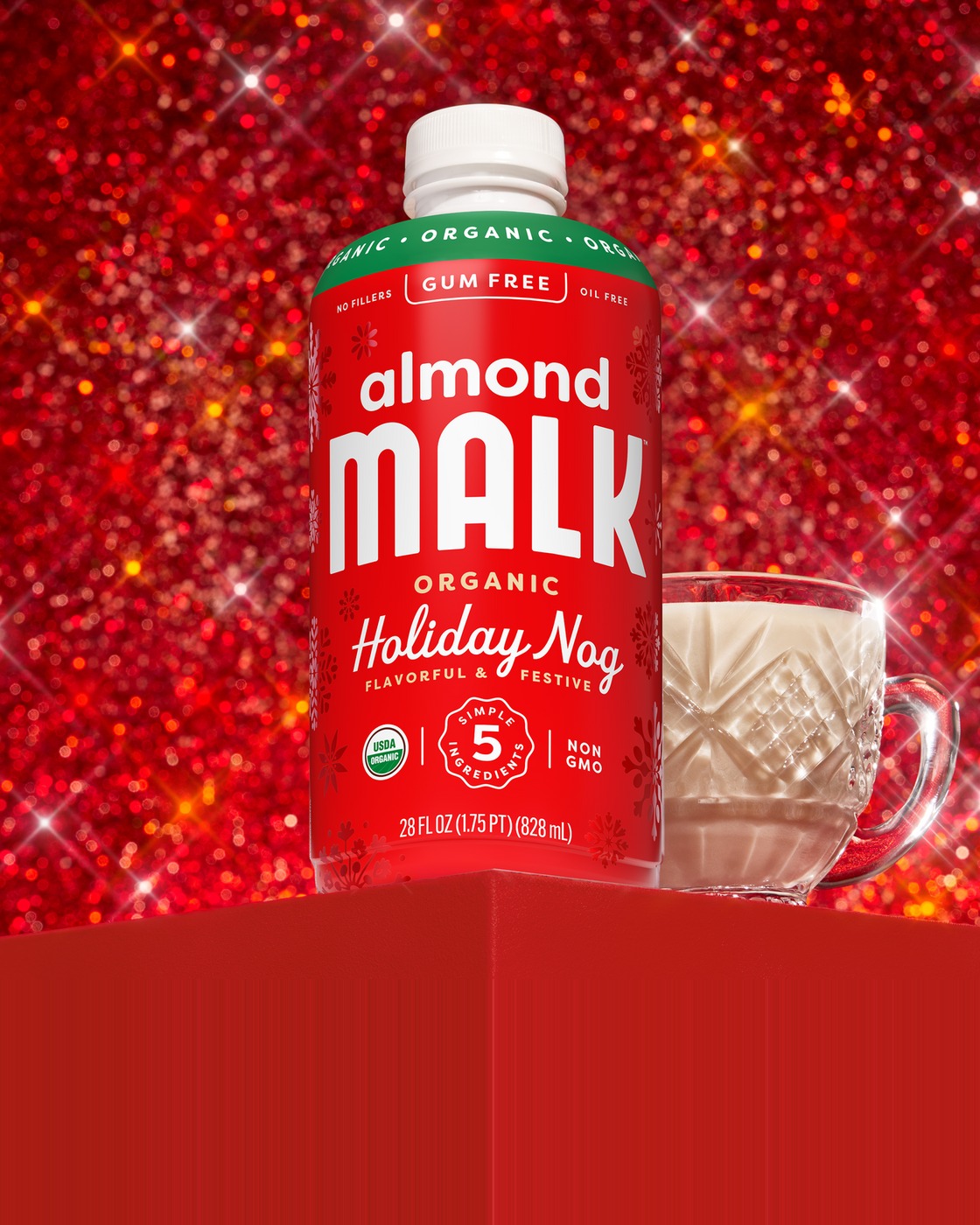 A bottle of MALK Holiday Nog Almond Milk beside a cut-glass cup on a red glitter backdrop.