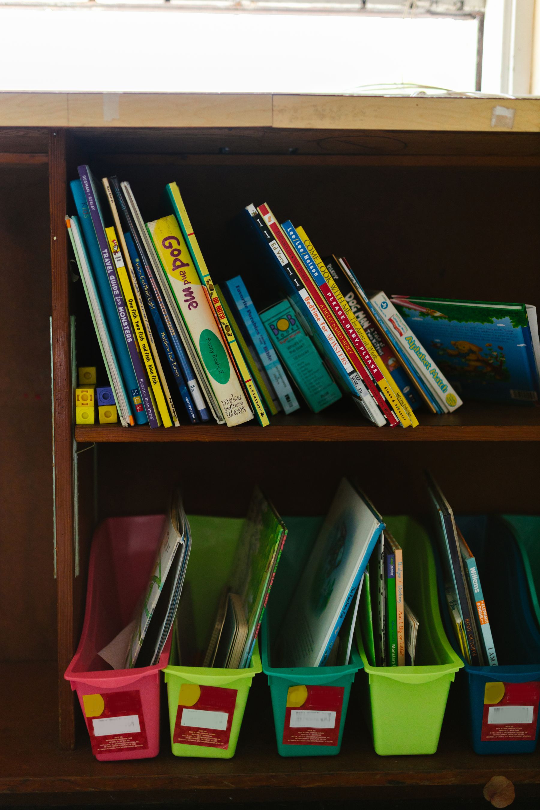 A low bookshelf of picture books with colored bins of books underneath.