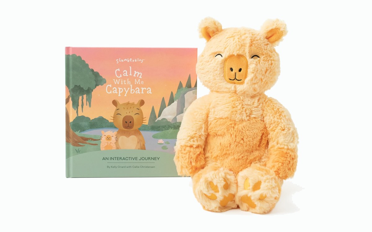 A coral weighted capybara plush sitting beside the hardcover book Calm With Me, Capybara, on a white background.