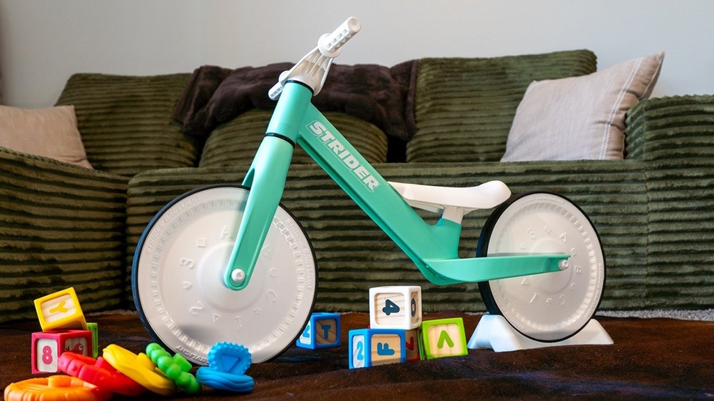 The green Strider Baby Balance Bike standing on a floor beside wooden alphabet blocks and teething toys.