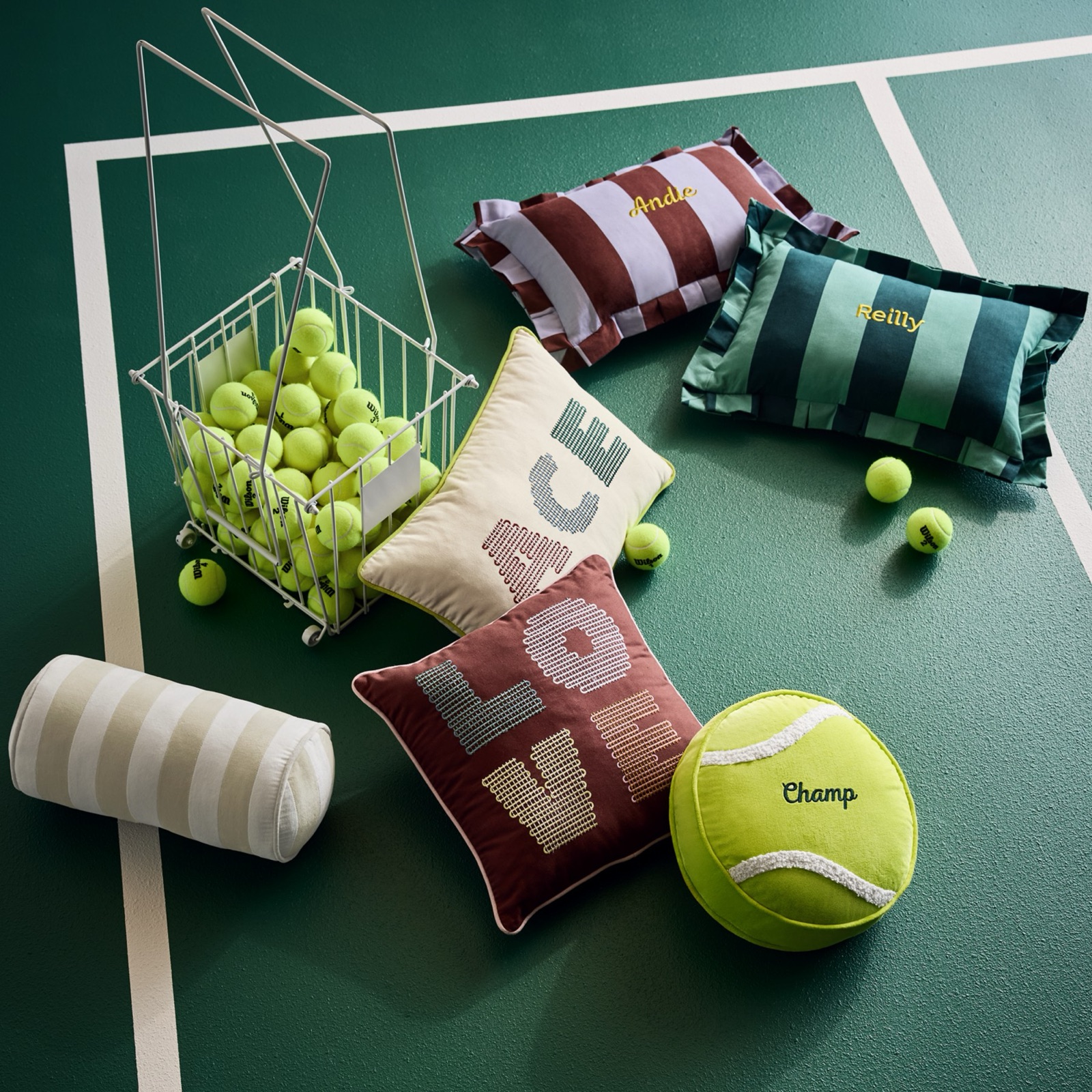 Striped pillow shams embroidered with names, a burgundy Love pillow and a tennis-ball pillow laid out on a green tennis court beside a basket of balls.