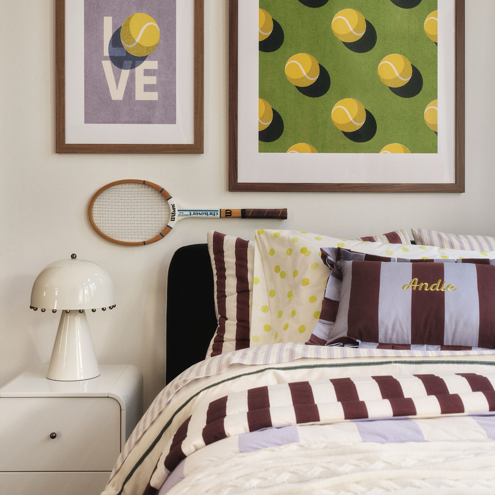 A kids bed made up in lavender and burgundy stripes, under two framed tennis-ball prints and a wooden racket hung on the wall.